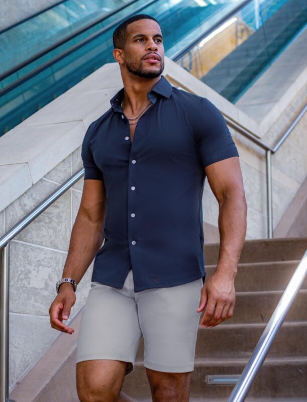 A man walks down an outdoor staircase wearing a navy blue State & Liberty short-sleeve button-down shirt paired with light grey stretch performance shorts. The athletic-fit shirt is tailored to the torso with white buttons, and the look is accessorized with a silver watch and chain. The background features modern stone stairs and a glass-paneled escalator.