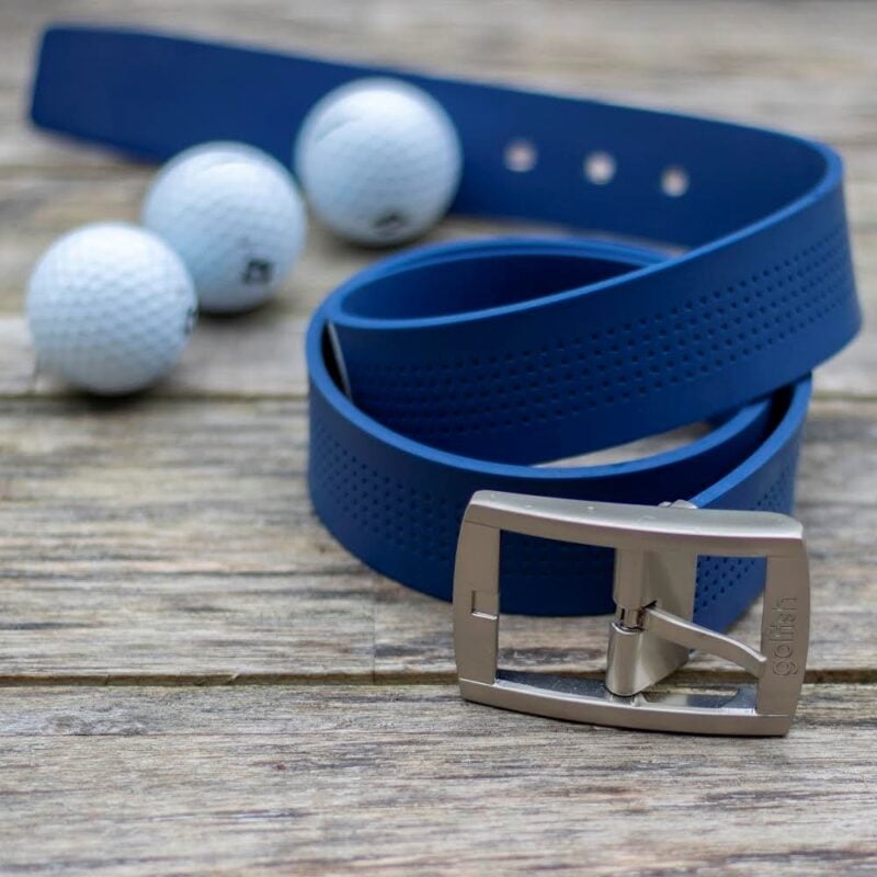 A navy blue Golfish brand golf belt is coiled on a rustic wooden surface next to three white golf balls. The belt features a perforated texture and a brushed silver buckle engraved with the "golfish" logo. The background is softly blurred, focusing on the detail of the belt's material and hardware.