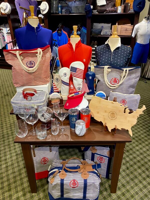 A patriotic merchandise display at Loblolly Golf Course in Hobe Sound, FL, featuring red, white, and blue apparel and accessories. The wooden table showcases striped tote bags, star-patterned shirts, golf headcovers with the American flag, and various glassware including wine and martini glasses. A large wooden cutting board shaped like the United States sits in the foreground, and all items feature the Loblolly pine tree logo.