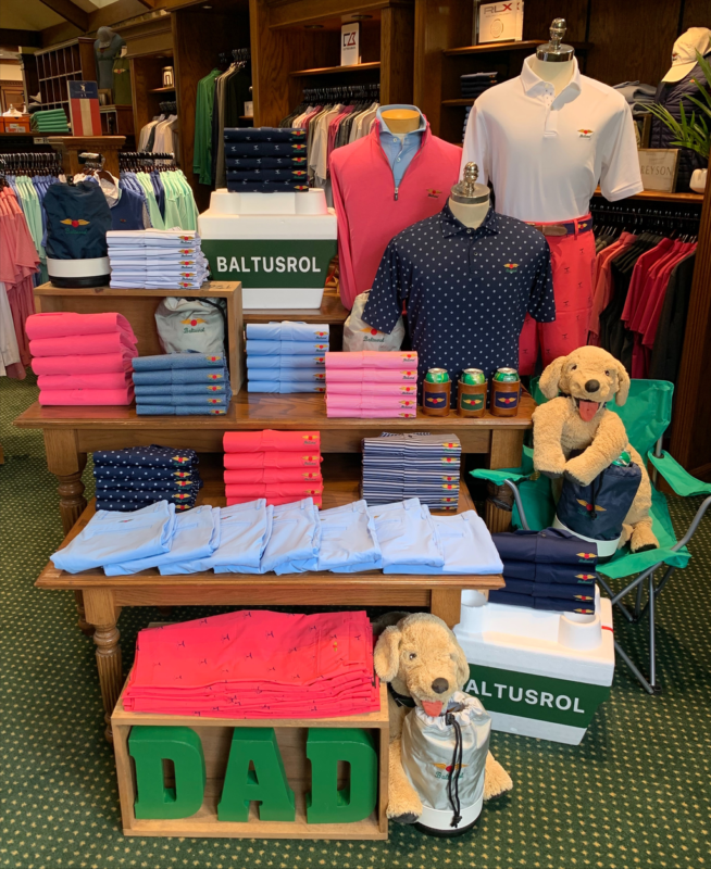 A Father's Day retail display at Baltusrol Golf Club featuring a "DAD" theme in green block letters. The wooden tables are organized with stacks of performance polos and shorts in vibrant pink, light blue, navy, and white, all featuring the club's winged logo. The display includes white and green "BALTUSROL" branded coolers, leather-wrapped canned beverage holders, and two plush golden retriever dogs posed with golf accessory bags.