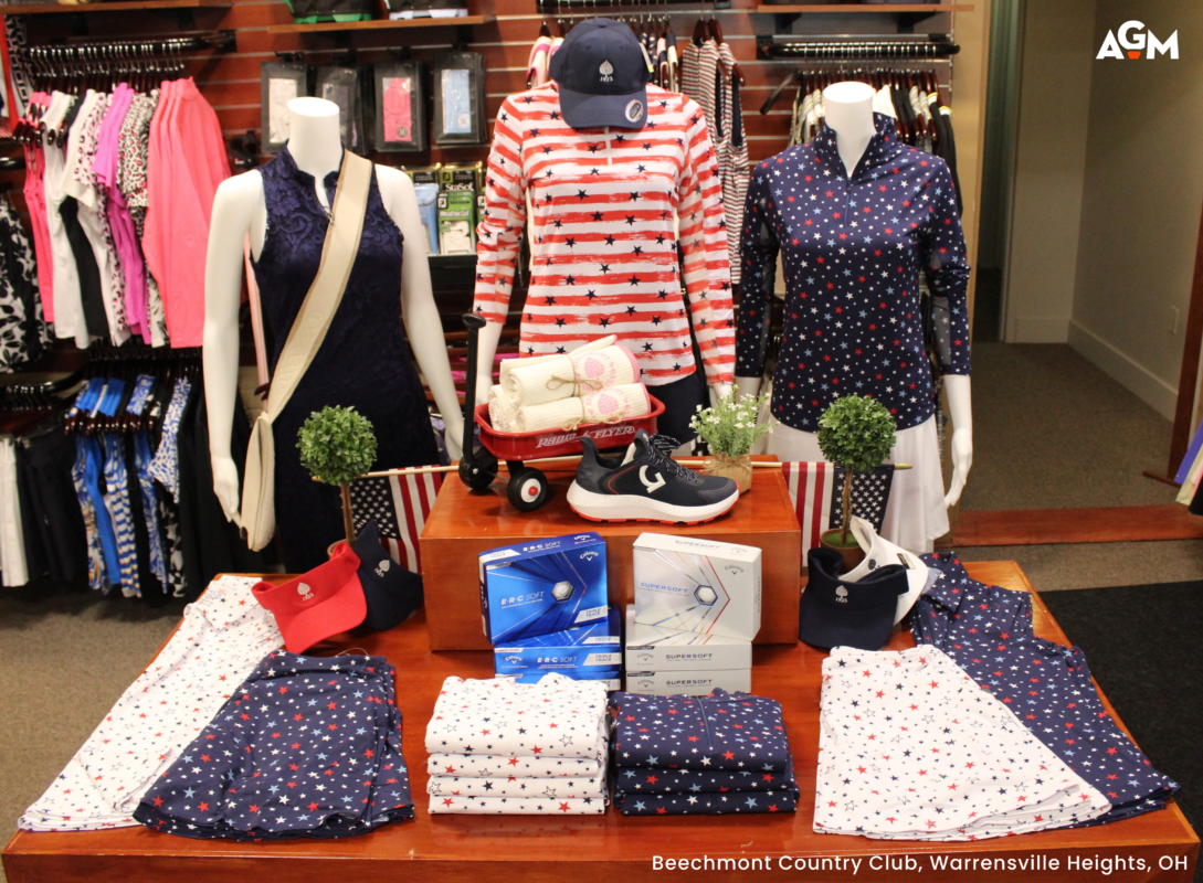 A Fourth of July women’s golf apparel display at Beechmont Country Club in Warrensville Heights, OH. Three mannequins showcase patriotic styles, including a navy lace dress, a red and white striped long-sleeve star print top, and a navy star-patterned polo. The central wooden table features a miniature red Radio Flyer wagon holding rolled towels, Callaway golf ball boxes, and folded star-print skorts in red, white, and blue.
