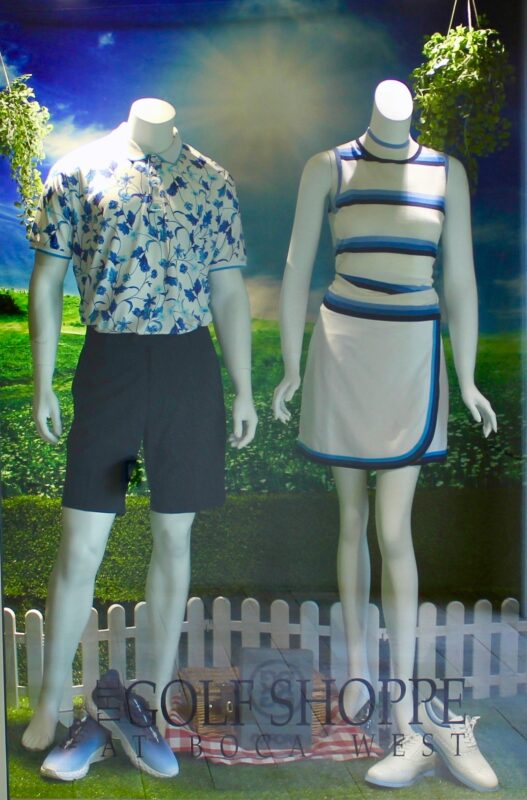 A "Golf Shoppe at Boca West" window display featuring two mannequins in blue and white golf attire against a scenic countryside backdrop. The male mannequin wears a blue floral print polo with navy shorts, while the female mannequin wears a striped sleeveless top and a white wrap skort with blue trim. The display is set on artificial turf with a white picket fence, a picnic basket on a checkered cloth, and hanging greenery.