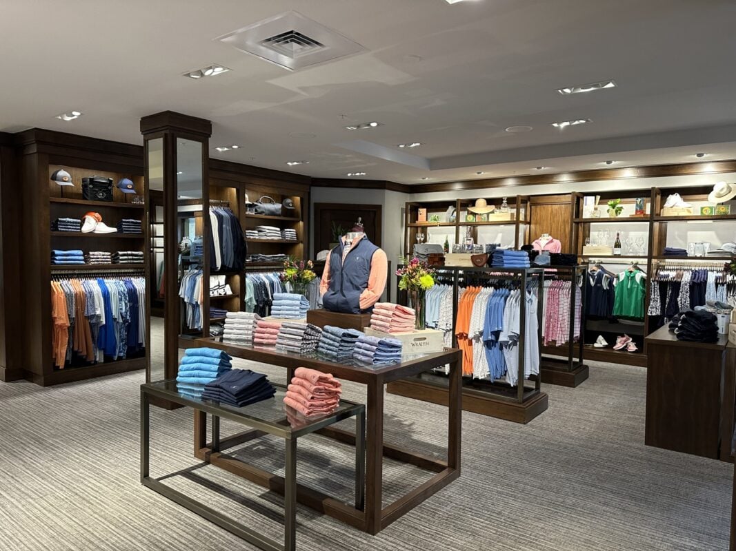A spacious, well-lit golf pro shop at Castle Pines Golf Club, featuring dark wood built-in shelving units filled with various apparel and accessories. Centered in the room are tiered wooden display tables showcasing neatly folded shirts in shades of blue and coral. The shop has neutral grey carpeting and a modern mannequin in the center wearing a navy vest and coral long-sleeve shirt.