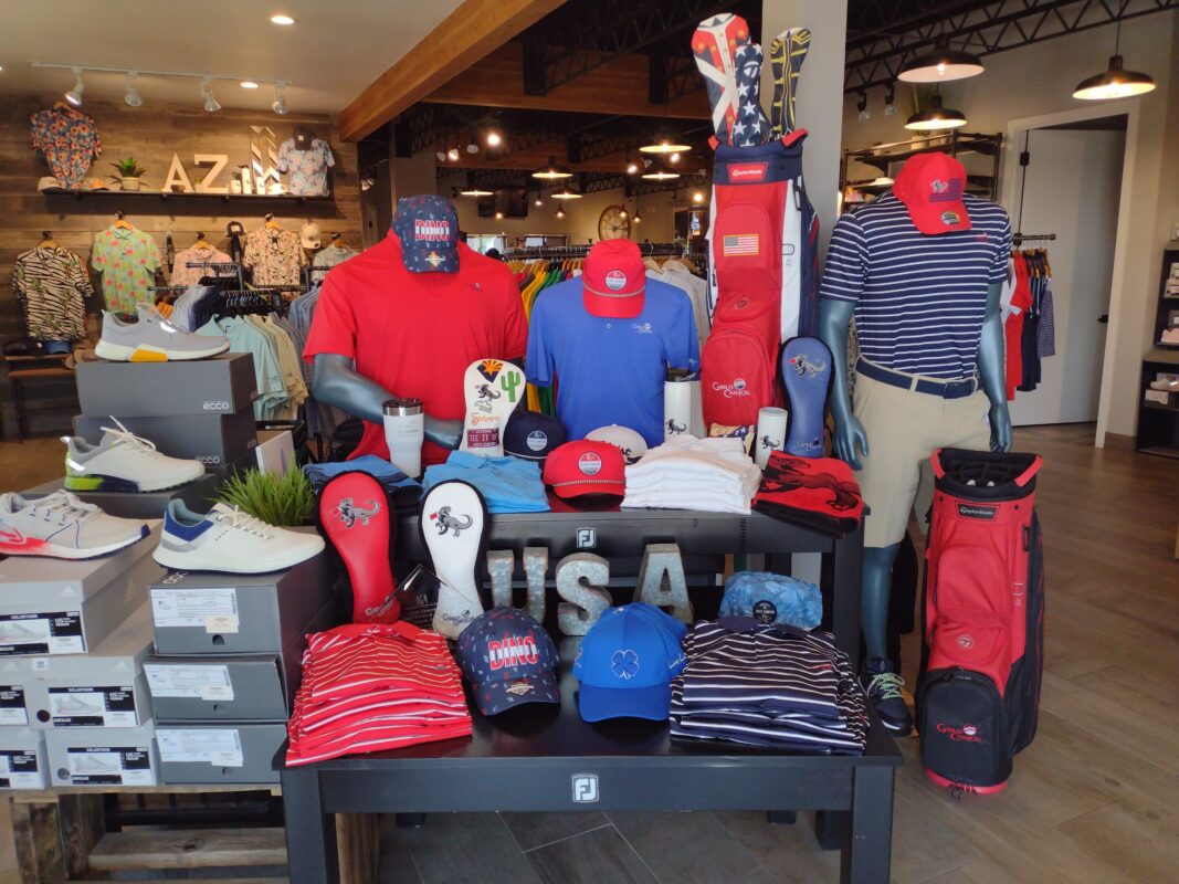 A Fourth of July-themed golf apparel display at Gold Canyon Golf Resort in Gold Canyon, AZ. A black table features large "USA" block letters, red and blue polo shirts, "DINO" branded hats, and various golf headcovers featuring a dinosaur logo and cactus embroidery. Red TaylorMade golf bags are positioned alongside mannequins wearing Americana-styled apparel, while boxes of ECCO golf shoes are stacked to the left.