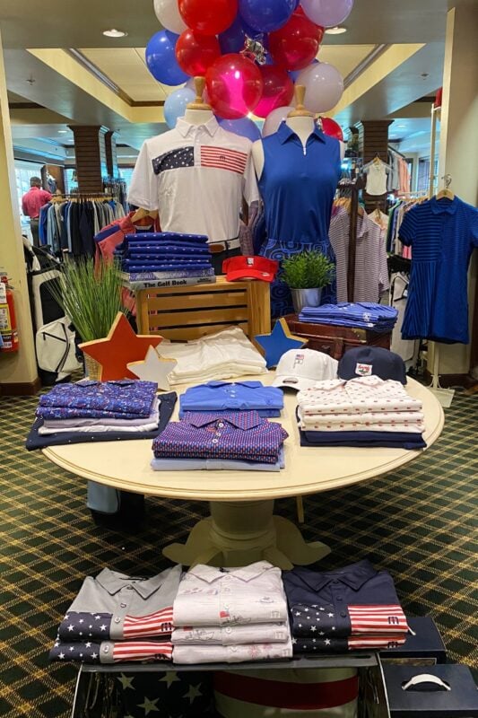A retail display with a patriotic American theme, featuring red, white, and blue golf apparel and decorations. Two mannequins stand at the back; one wears a white polo with a stars-and-stripes chest graphic, and the other wears a solid royal blue sleeveless top. Red, white, and blue balloons are clustered above them. A round white table in the foreground showcases neatly folded shirts in various patterns, including stars and stripes, along with white and navy golf caps and decorative wooden stars.