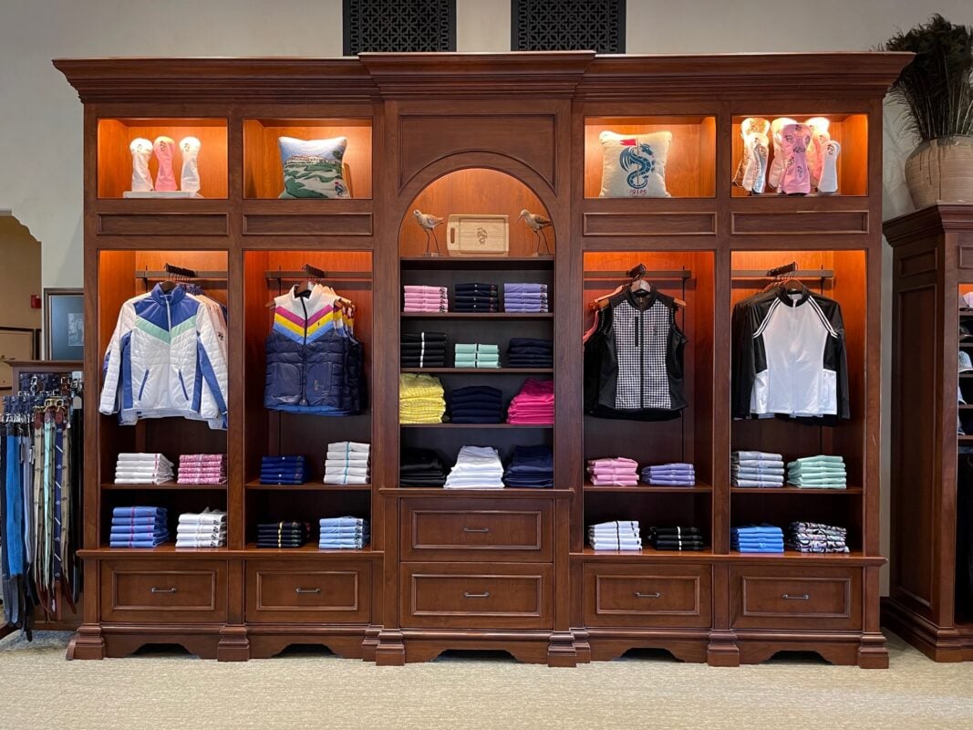 A large, ornate dark wood wall unit at Monterey Peninsula CC featuring a dedicated women's apparel display. The lit shelving sections showcase various folded shirts in pink, blue, and white, alongside hung vests and jackets. Decorative elements include embroidered golf pillows and bird figurines in a central arched alcove.