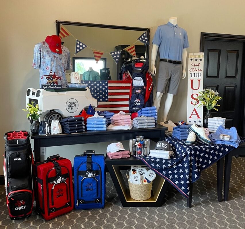 A patriotic July 4th retail display at Oaks Country Club featuring a "God Bless the USA" theme. The setup includes red and blue "OAKS" branded rolling luggage, a large white cooler with an oak leaf logo, and Sun Mountain golf bags. Mannequins model light blue apparel against a backdrop of American flag bunting and a wooden flag sign, while star-patterned blankets and folded polos in shades of red, white, and blue are arranged across black display tables.