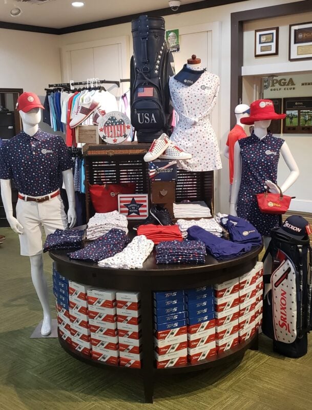 An elaborate USA-themed merchandise display at PGA Golf Club featuring star-patterned apparel and accessories. A circular tiered table is stocked with red, white, and blue golf ball boxes (TaylorMade TP5 and Titleist) arranged to mimic stripes. Mannequins model navy and white star-print polos and dresses, complemented by a navy "USA" golf bag, red headwear, and themed footwear.
