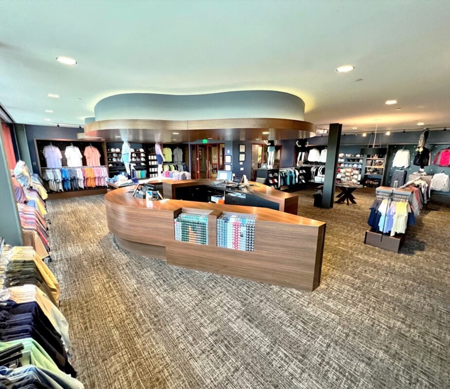 A wide-angle view of the modern golf pro shop at Streamsong Resort, featuring a large, curved wooden service counter as the center focal point. The shop has dark blue walls, grey patterned carpeting, and sleek recessed lighting, with various racks and wall displays showcasing a wide range of colorful golf apparel and accessories.