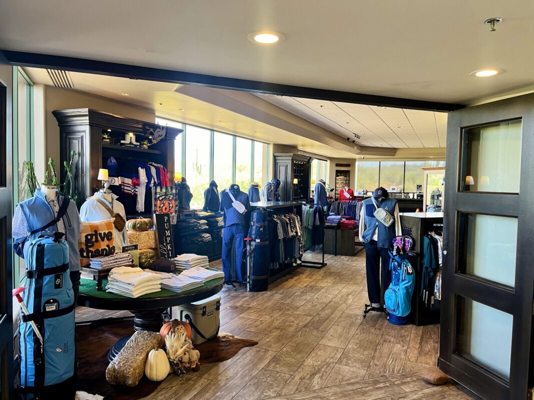 A spacious, light-filled golf pro shop at Troon North with large windows overlooking a bright landscape. In the foreground, a round display table features folded white shirts, a "give thanks" pillow, and fall-themed decor like pumpkins and hay bales. To the left, a mannequin in a light blue vest stands next to a matching blue golf bag, while additional apparel racks and dark wood shelving units fill the rest of the shop.