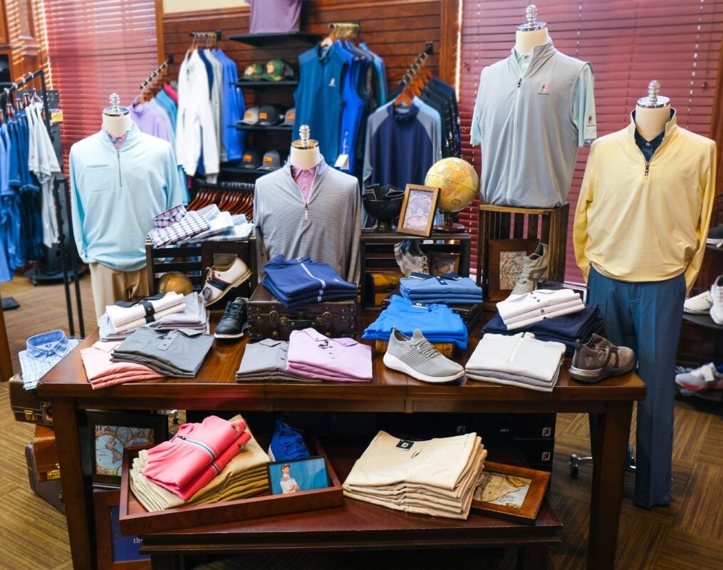 A well-organized display of golf apparel at Terradyne Country Club in Andover, KS. The foreground features a dark wood table stacked with neatly folded polo shirts in pink, grey, blue, and white, alongside casual grey athletic shoes. Four mannequins are dressed in various quarter-zip pullovers and vests in light blue, striped grey, and pale yellow. The background includes additional clothing racks, a globe, and framed artwork, all set against rich wood-paneled walls and blinds.