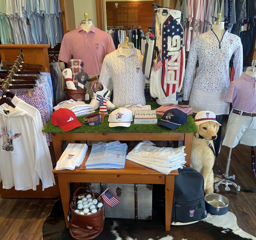 A Fourth of July retail display at Washington Golf & Country Club featuring patriotic apparel and accessories. A wooden table topped with green turf showcases red, white, and blue polos, RLX caps featuring a golfing bear, and a white PING golf bag with American flag stars and stripes. Mannequins model striped and patterned golf attire, while a life-sized golden retriever statue wearing a club-branded bucket hat sits beside a leather bag filled with golf balls and small American flags.