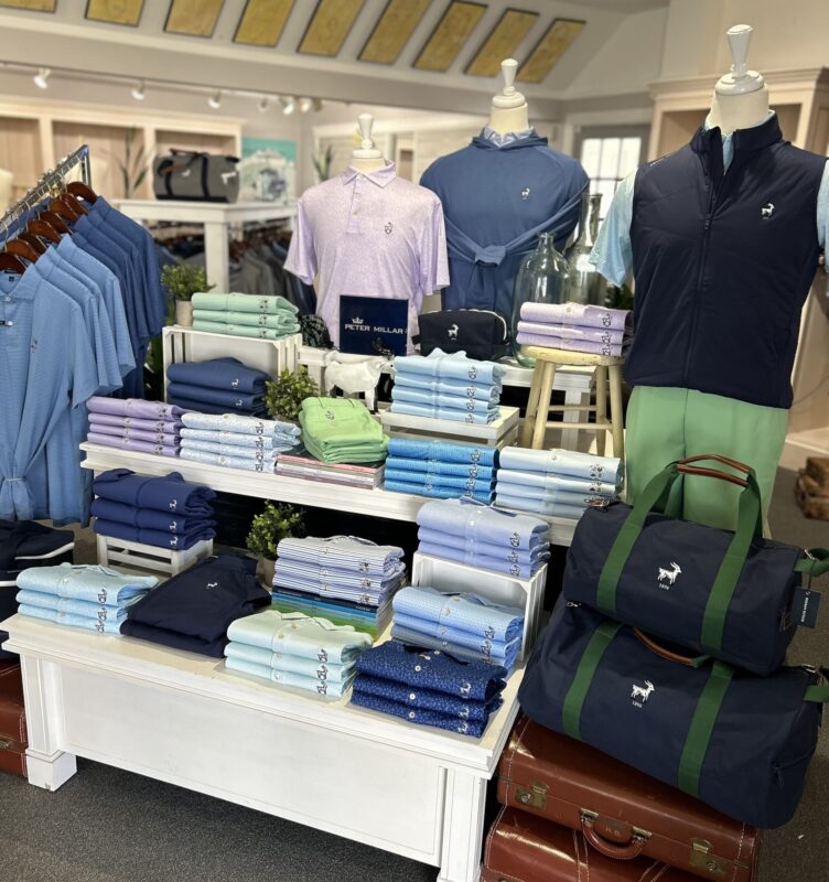 A retail display at Wannamoisett CC featuring Peter Millar apparel. Neatly folded polos in various shades of blue, purple, and green are arranged on tiered white tables and wooden crates. Three mannequins showcase a light purple polo, a blue hoodie, and a navy vest with green trousers. Two navy duffel bags with green straps are stacked on vintage brown suitcases in the foreground.