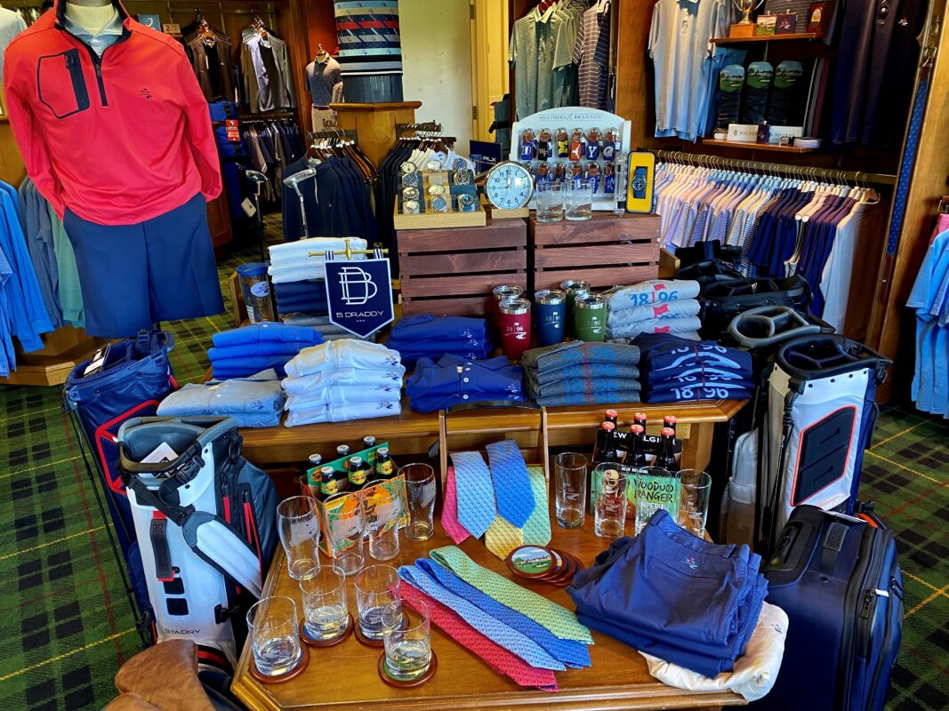 A Father's Day retail display at Wee Burn Country Club in Darien, CT, featuring various luxury gifts and apparel. A wooden table is styled with stacks of B. Draddy performance polos, vibrant patterned neckties, leather-strapped watches, and various glassware including pint and rocks glasses. The setup is anchored by white and navy Titleist StaDry golf bags and includes a display of Smathers & Branson needlepoint key fobs and a New Belgium Voodoo Ranger beer pack.