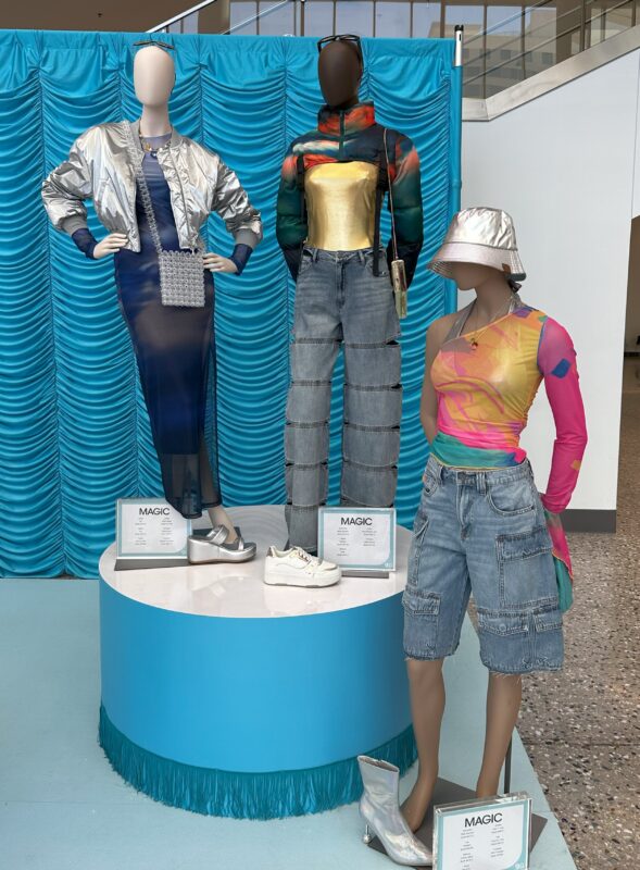 Three mannequins styled in colorful, contemporary outfits—including denim, cropped tops, and a bucket hat—displayed on a round platform at a MAGIC trade show booth with a blue backdrop.