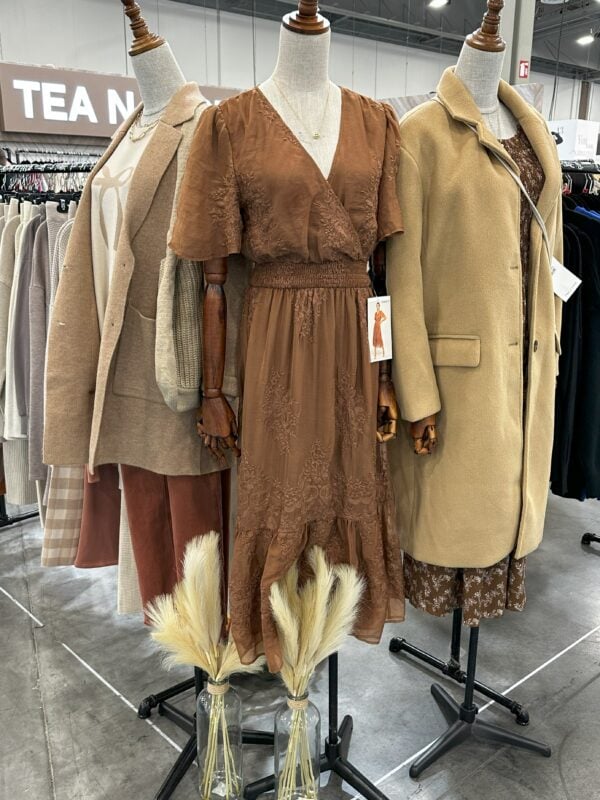 Three mannequins styled in neutral-toned women’s apparel, including a brown lace midi dress and layered tan outerwear, displayed at a trade show booth with dried floral accents in front.