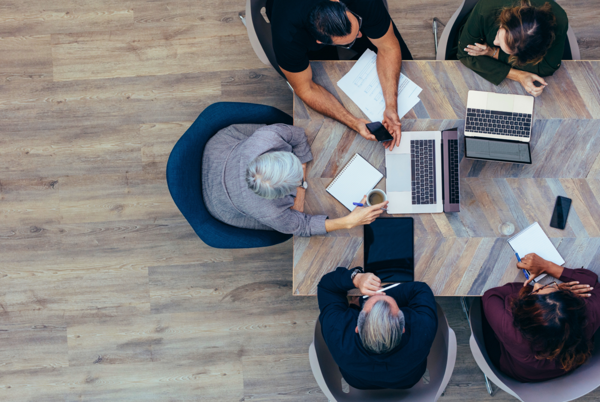 Overhead view of four colleagues seated around a wooden conference table with laptops, notebooks, smartphones, and coffee, collaborating during a meeting.