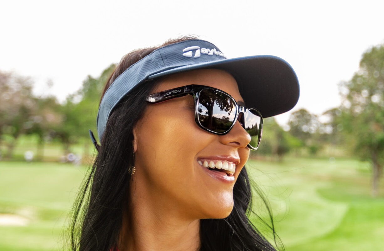 Woman wearing black JONDO sunglasses and a blue TaylorMade visor, smiling on a golf course with trees and fairway in the background.