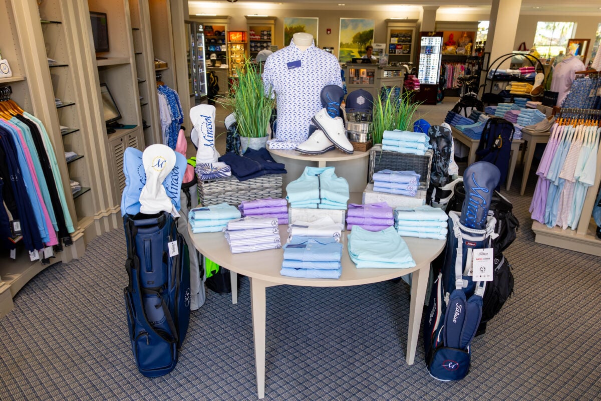 Golf shop display at The Country Club at Mirasol featuring a round table neatly stacked with folded polos in pastel shades of blue, purple, and aqua. A central mannequin displays a patterned shirt alongside white golf shoes and hats. Branded "Mirasol" headcovers top nearby golf bags, with apparel and accessories arranged on surrounding shelves and racks.
