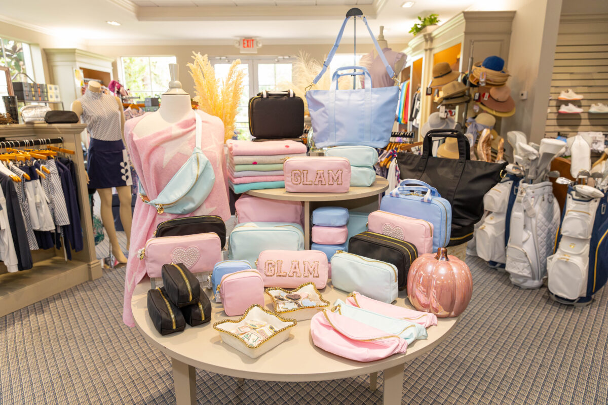 A retail display at the Country Club at Mirasol featuring a variety of Stoney Clover Lane accessories in a pastel "candy" color palette of soft pink, mint green, and sky blue. A round, two-tiered wooden table is meticulously arranged with zippered pouches of various sizes, some featuring "GLAM" in gold glitter chenille patches and others with pearl-beaded hearts. The display includes a mannequin draped in a pink star-patterned wrap wearing a mint belt bag, several large tote bags, and decorative accents like a metallic rose gold pumpkin and gold-rimmed trinket dishes. In the background, white and navy golf bags and additional women's golf apparel are visible in the brightly lit pro shop.