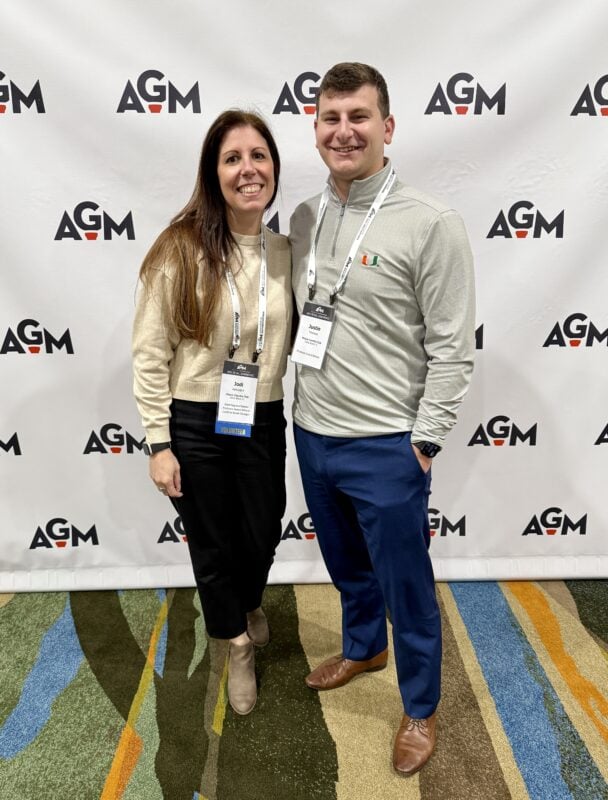 A smiling woman and man stand in front of an AGM step-and-repeat backdrop at a professional event. Both wear conference name badges and lanyards. The woman has long brown hair and is dressed in a cream sweater, black pants, and taupe ankle boots. The man wears a light gray pullover with the University of Miami logo, blue pants, and brown dress shoes. They are standing on a multicolored carpet with blue, green, orange, and tan accents.