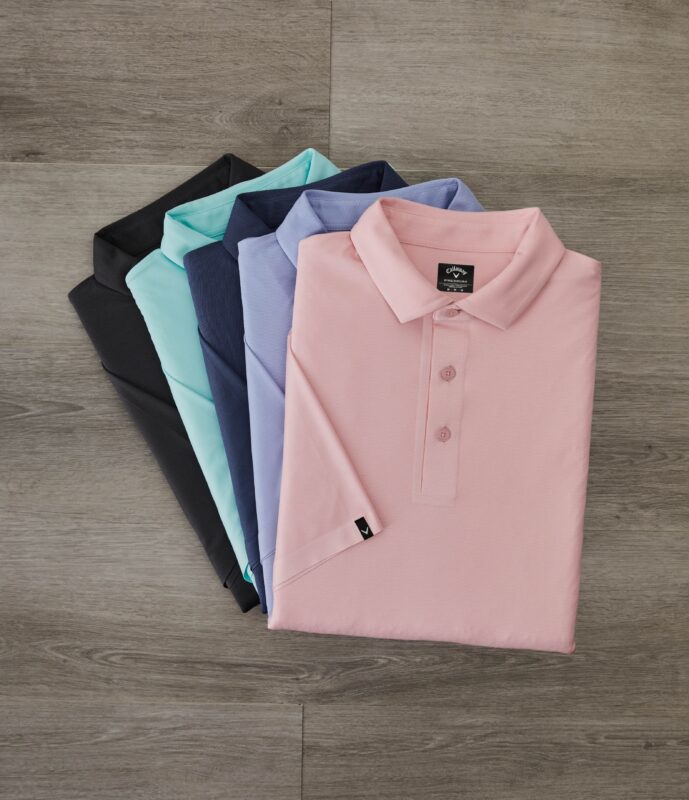 Flat lay of six neatly folded Callaway golf polos arranged in a fan shape on a gray wood floor. The shirts are displayed in a gradient from black, teal, navy, lavender, and periwinkle to light pink, with the pink polo on top showing a visible Callaway logo on the label and sleeve tag.