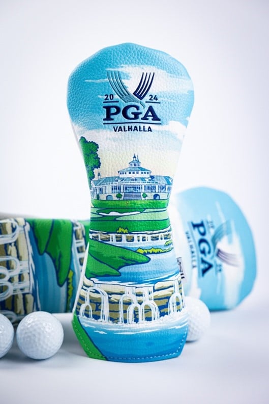 A 2024 PGA Championship headcover designed for Valhalla Golf Club, featuring colorful illustrations of the clubhouse, course landscape, and iconic bridges. The headcover has a bright blue sky background and sits upright among a few white golf balls, with additional headcovers blurred in the background.