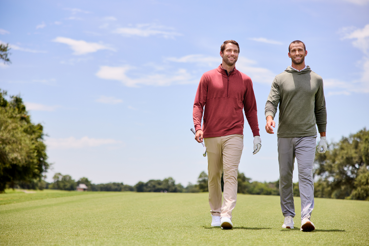 Two men walk side by side on a sunny golf course, smiling and wearing golf gloves. One wears a brick red quarter-zip pullover and beige pants, while the other wears an army green hoodie and gray pants. Both hold golf clubs, with trees and a blue sky in the background.