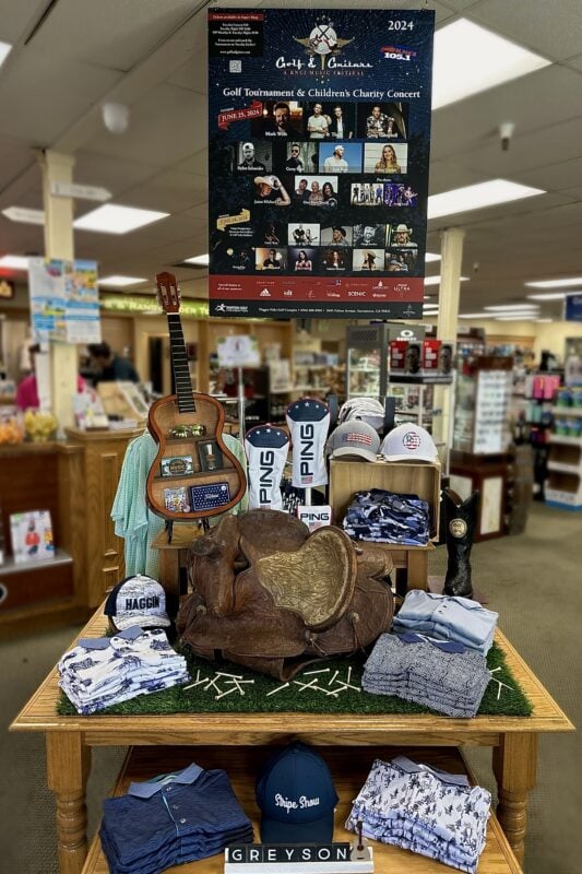 In-store golf shop display featuring a rustic western theme with a vintage leather saddle, wooden guitar shadowbox, cowboy boot, and stacked men's golf apparel in blue and white tones. Above the table is a promotional poster for the 2024 "Golf & Guitars" charity tournament and concert on June 25. Branded items from PING, Haggin Oaks, and Greyson are visible throughout the setup.