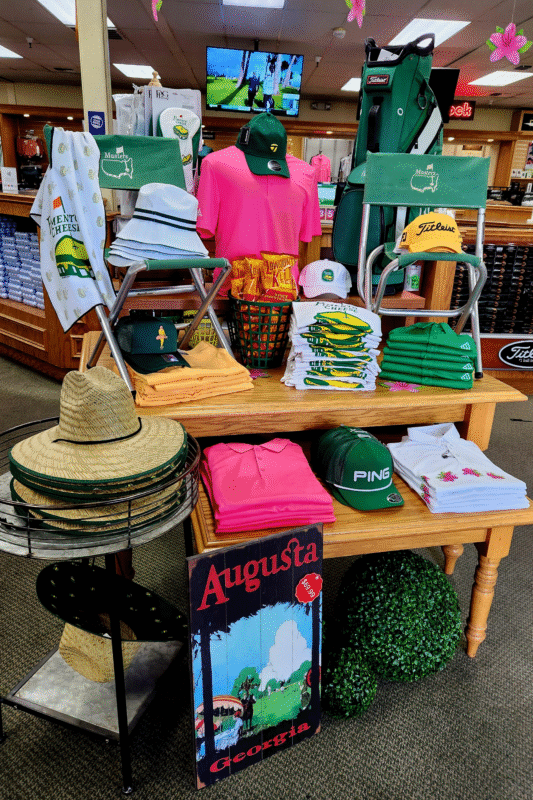 Augusta Tournament-themed golf shop display featuring vibrant green, yellow, and pink merchandise, including folding chairs, shirts, hats, and towels with Masters logos. A basket of Moon Pies, a Titleist golf bag, and an "Augusta, Georgia" wooden sign enhance the Augusta National theme. Straw hats and azalea decorations complete the festive and iconic tournament atmosphere.
