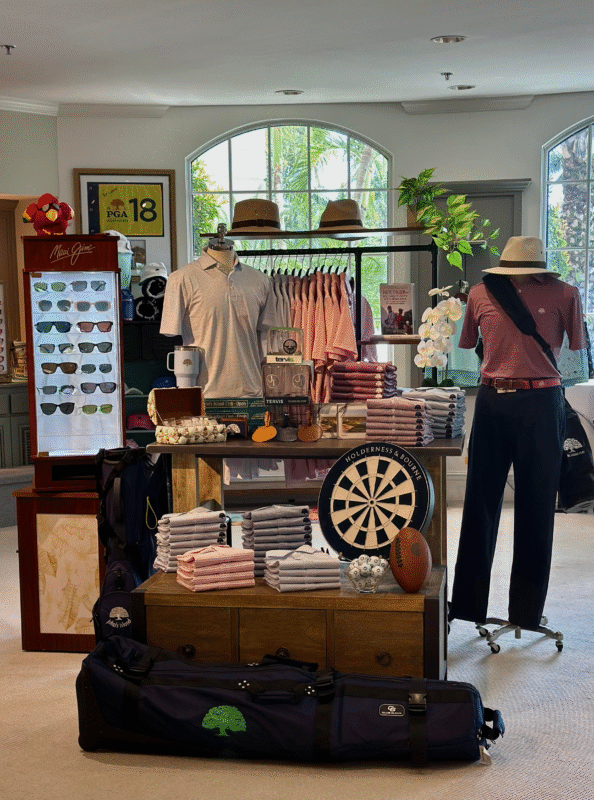 A stylish golf shop display at John’s Island Club featuring neatly folded polos in soft pink, gray, and lavender tones, arranged around mannequins dressed in coordinating outfits. A dartboard labeled "Holderness & Bourne," golf accessories, sunglasses, Tervis tumblers, and travel bags are used as themed props. Tropical plants and natural light from large arched windows add a fresh, upscale atmosphere.