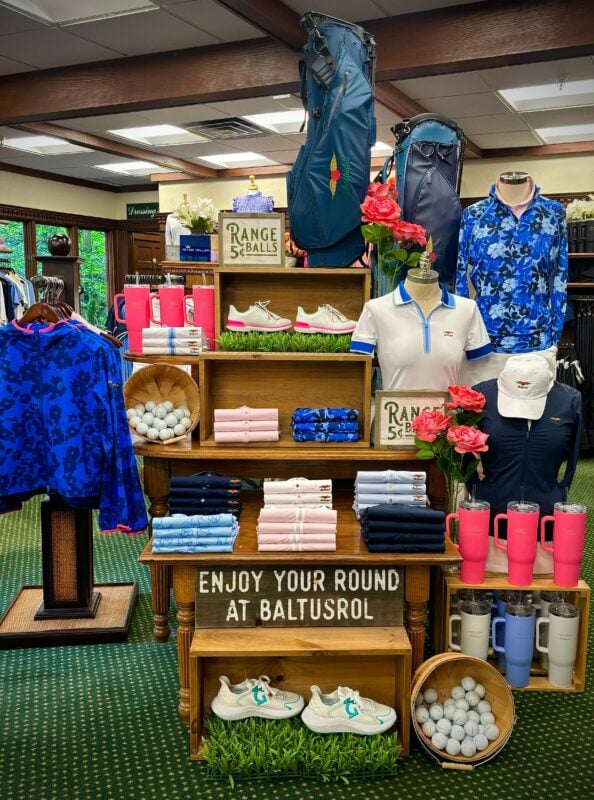 Eye-catching golf shop display at Baltusrol Golf Club featuring a variety of folded polos, jackets, golf shoes, tumblers, and accessories in coordinated pink, blue, and white tones. Two golf bags stand at the back with a “Range Balls 5¢” sign and artificial grass, flowers, and baskets of golf balls used as decorative accents. A wooden sign at the bottom reads, “Enjoy your round at Baltusrol.”