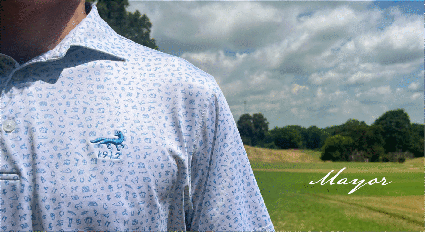 Close-up of a person wearing a white collared polo shirt with a light blue all-over print featuring small icons. The shirt has an embroidered blue fox logo above the number "1912" on the left chest. The background shows a grassy field with trees and a partly cloudy sky.