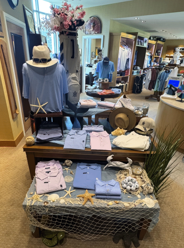 Coastal-themed golf shop display at Belmont Country Club featuring pastel men’s polos in blue, lavender, and pink, decorated with starfish, seashells, netting, and faux sea grass. A mannequin in a light blue shirt and straw hat stands beside a golf bag adorned with flowers. The setup evokes a beachy, nautical vibe while showcasing spring and summer apparel.