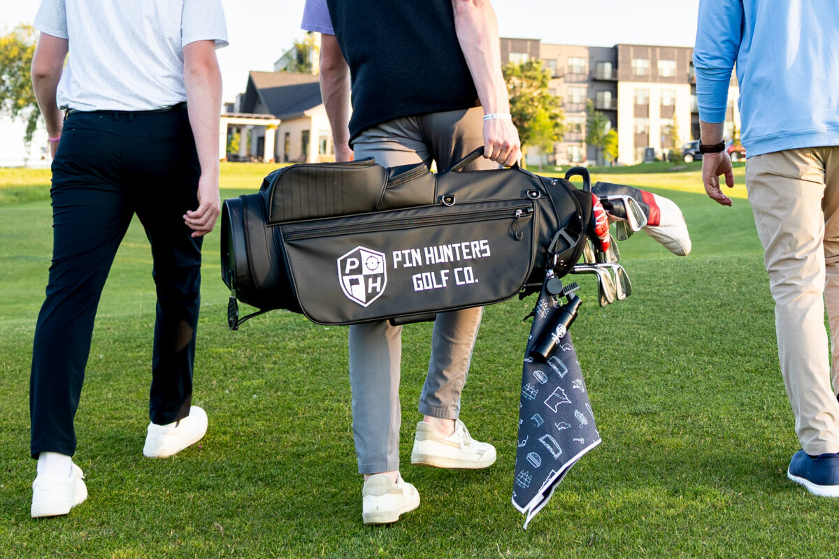 Three men walk across a golf course, with the center person carrying a black golf bag that reads "Pin Hunters Golf Co." in white text. A dark patterned golf towel with white graphics hangs from the bag, and golf clubs are visible inside. Buildings and trees are seen in the background.