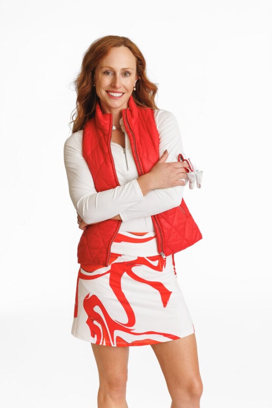 Woman smiling in a studio setting, wearing a red quilted vest over a white long-sleeve top and a white golf skirt with bold red abstract patterns. She holds a golf glove in one hand and exudes a confident, sporty look.