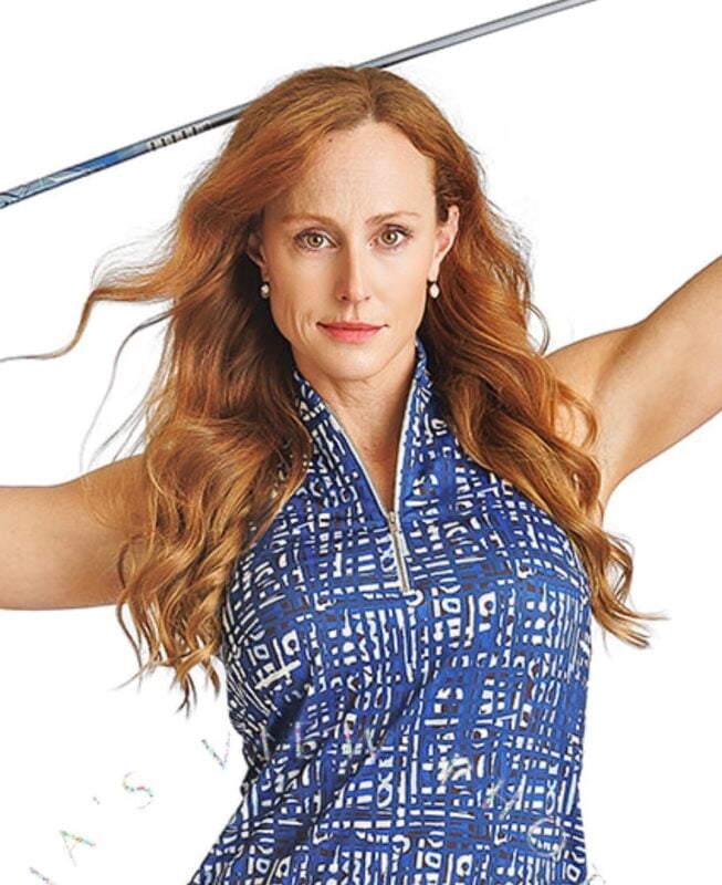 Woman with long wavy red hair holds a golf club behind her shoulders while wearing a sleeveless zip-collar top with a bold blue and white geometric pattern, posing confidently against a white backdrop.
