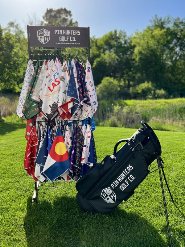A display rack showcasing a colorful variety of printed golf towels from Pin Hunters Golf Co., arranged in two rows on hangers outdoors on a sunny day. The sign above the rack features the brand’s logo and website. A sleek black Pin Hunters Golf Co. bag with white branding rests on a stand beside the display, all set against a vibrant green lawn and tree-filled background.