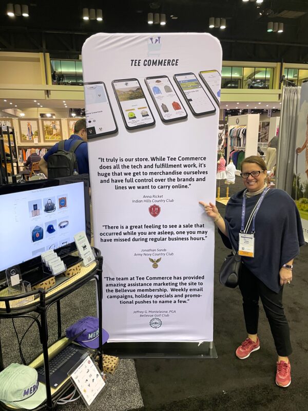 Woman standing beside a Tee Commerce trade show display featuring a large banner with customer testimonials and images of mobile online shops, pointing to the sign while smiling, with apparel booths visible in the background.
