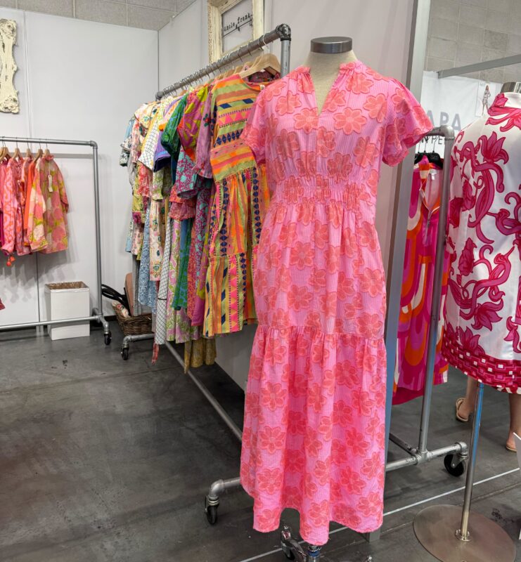 A mannequin displays a pink and coral floral maxi dress with a gathered waist in a colorful boutique-style booth. Surrounding racks showcase vibrant, patterned dresses in bright hues like pink, yellow, and green, creating a cheerful, summery atmosphere.