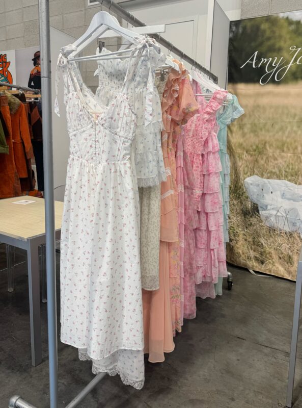 A clothing rack displays a collection of romantic, vintage-inspired dresses in soft pastel colors and floral prints at a boutique trade show. The dresses feature details like ruffles, lace trim, smocking, and tie straps, evoking a cottagecore aesthetic.