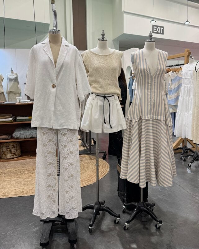 Three mannequins display neutral-toned women's outfits with a breezy, minimalist aesthetic at a boutique fashion trade show. The left mannequin wears a white linen blazer over sheer lace pants. The center mannequin is dressed in a cream knit top paired with white shorts featuring a black drawstring. The right mannequin showcases a sleeveless beige-and-white striped dress with a plunging neckline and flared hem. The backdrop includes woven rugs and a rustic wooden shelving unit with neatly folded garments.