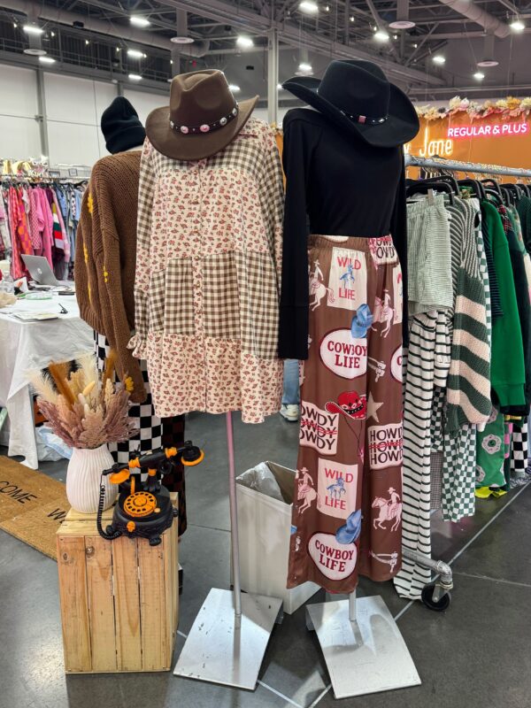 Two mannequins showcase Western-inspired outfits at a boutique trade show. One wears a brown cowboy hat and patchwork dress with floral and gingham prints, while the other wears a black cowboy hat, black top, and wide-leg pants printed with cowboy boots, hats, and phrases like "Howdy" and "Cowboy Life." A rustic wooden crate with a vintage toy phone and pampas grass decorates the booth.