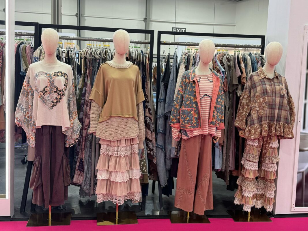 Four mannequins dressed in boho-inspired layered outfits with earth-toned tops, ruffled lace skirts, wide-leg pants, and printed jackets are displayed in front of a clothing rack at a trade show booth. The setting features a bright pink carpet and industrial ceiling lights overhead.