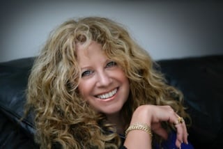 Jamie Sadock smiling and sitting on a black leather couch. She has long, curly blonde hair, is wearing a blue top and gold jewelry, including a bracelet and rings, and is looking directly at the camera.