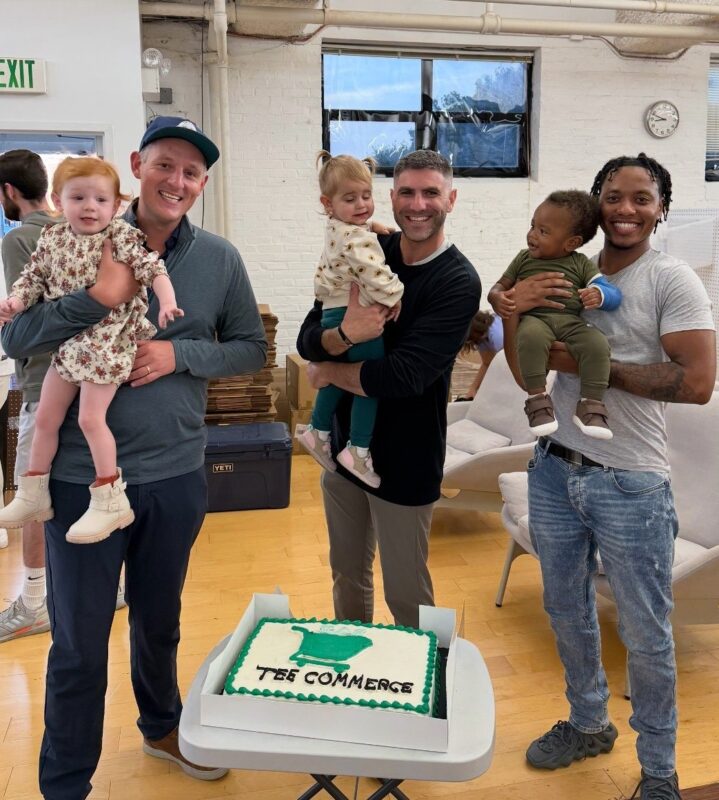 The kiddos of Tee Commerce: The photo depicts three smiling men each holding a young child in their arms, standing behind a cake that reads "Tee Commerce" with a green golf tee graphic. The casual indoor setting has white brick walls, a wood floor, and a laid-back, family-friendly atmosphere.