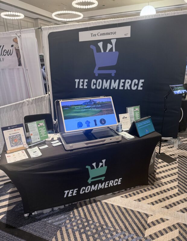 Tee Commerce trade show booth featuring a large branded backdrop and tablecloth with the company’s logo—a shopping cart filled with golf clubs. A laptop on the table displays a sample online golf shop for Sankaty Head Golf Club. Promotional materials, QR codes, and informational flyers are arranged on the table, with a sign above reading “Tee Commerce” and booth number 627.
