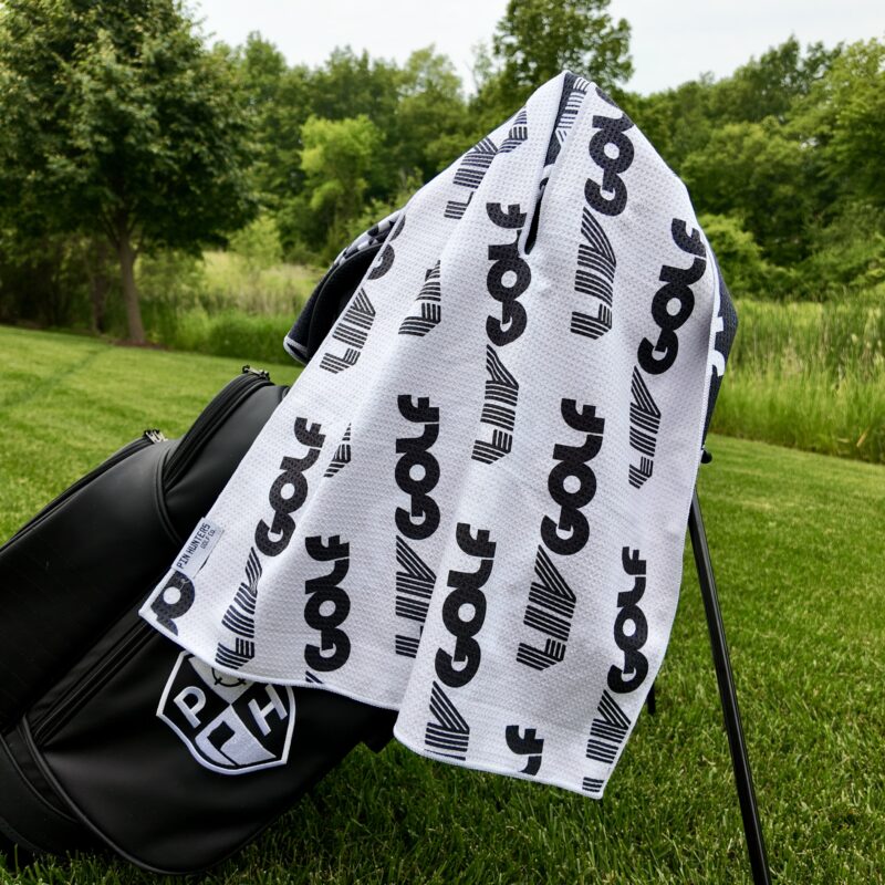 A black Pin Hunters Golf Co. bag sits on green grass with a white golf towel draped over it, featuring a bold black repeating pattern of the word “GOLF” and stylized “LIV” logos. A peaceful wooded area with trees and tall grass is visible in the background.