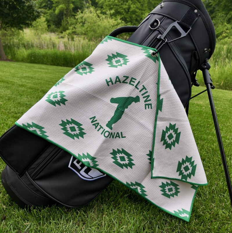 A black golf bag with a white and green towel draped over it, featuring the silhouette of a golfer and the words “Hazeltine National” in bold green letters. The towel is decorated with repeating geometric patterns and bordered in green, set against a grassy backdrop with trees and tall grass in the distance.