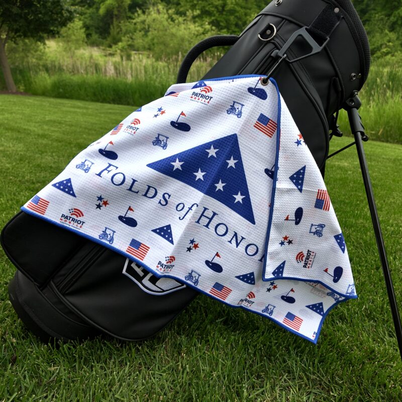 A black Pin Hunters Golf Co. bag stands on vibrant green grass with a patriotic golf towel draped over it. The towel features a navy blue triangle with stars and the text “Folds of Honor,” along with repeating American flag icons, golf carts, golf balls, and “Patriot Mobile” logos. A wooded area and tall grass are visible in the background.